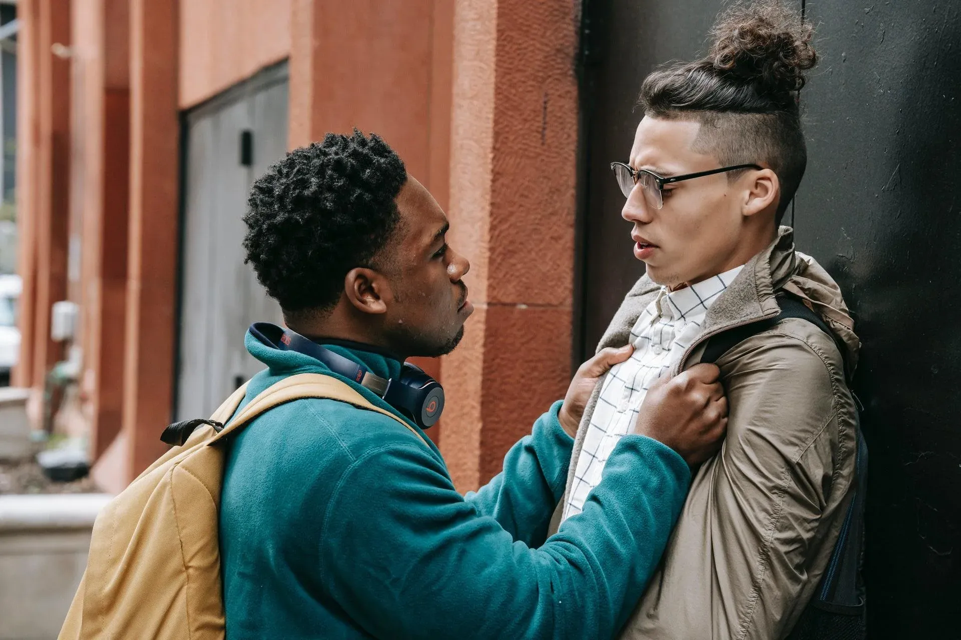 Lack of discipline for bullying is often one of the biggest reasons to homeschool according to surveyed parents. This image shows a boy wearing a backpack and headphones holding another boy up against a wall, gripping the front of his jacket. The boy on the wall looks frightened.